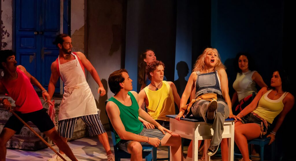 The ensemble sits together on chairs in warm light. Mamma Mia! (AG Theatre 2026), lighting design by ISLX