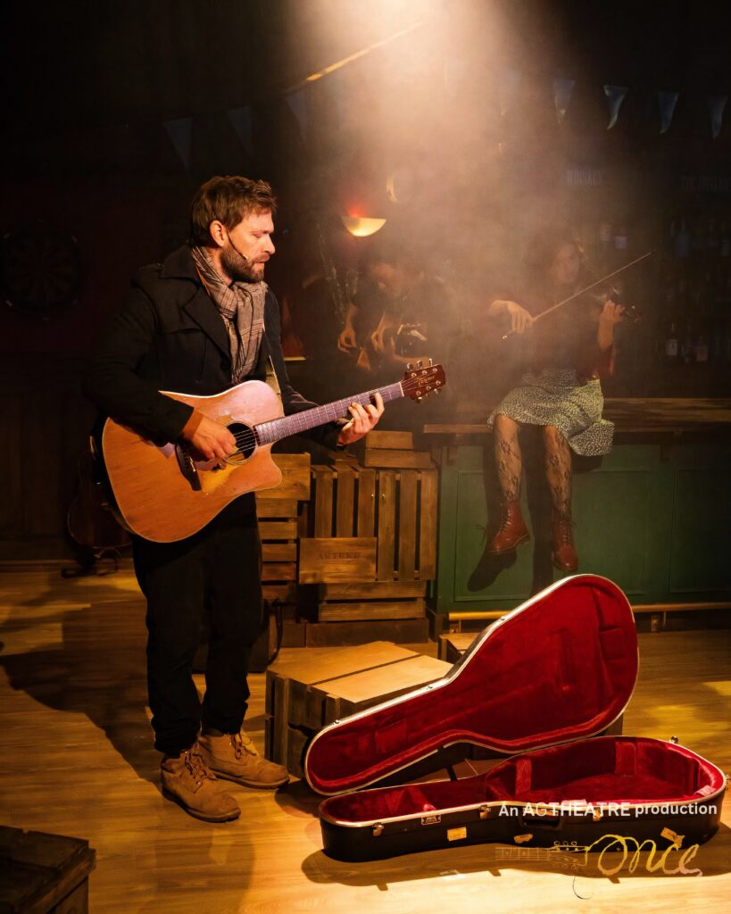 Our Work 2 Guy plays guitar in a shaft of light through the haze, guitar case open at his feet. Once (AG Theatre 2026), lighting design by ISLX