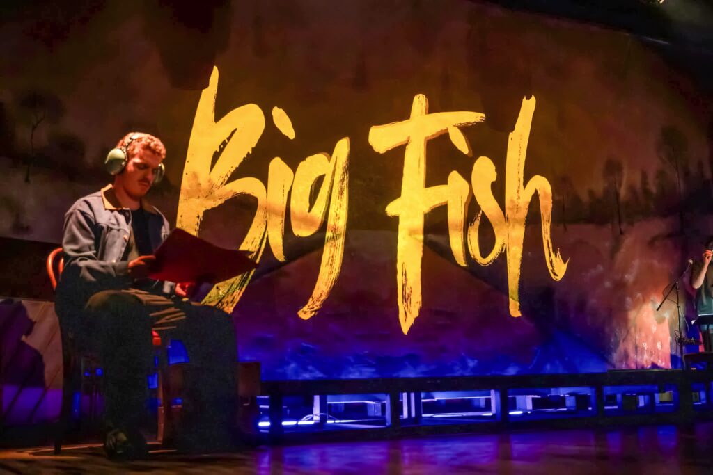 Our Work 4 Big Fish title projected in gold across the set as a performer reads. Big Fish (Western Arts Theatre 2026)